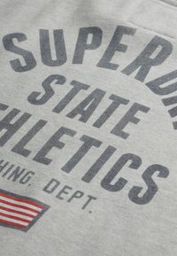 Tela gris con texto audaz en azul oscuro: "SUPERDRY STATE ATHLETICS", con un texto más pequeño debajo. Incluye un detalle de una bandera a rayas rojas y blancas.