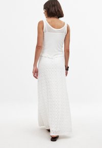 Jupe maxi blanche avec un motif texturé et des accents en dentelle, associée à un débardeur léger. Sandales plates et cheveux courts visibles.