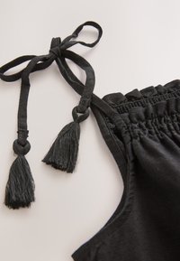 Black fabric close-up with a tied strap and tassel detailing. Features gathered elastic edging for adjustable fit. Smooth texture.