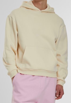 Persoon die een crèmekleurige hoodie met zak aan de voorkant en roze trainingsbroek draagt, staand met één hand in de zak tegen een effen achtergrond.