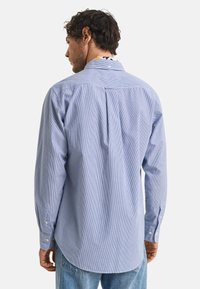 Camisa de manga larga a rayas en azul y blanco, con cuello clásico, puños con botón y un detalle de pliegue en la parte posterior.