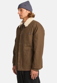 Brown canvas jacket with a cream sherpa collar, front button closure, and side pockets. Worn over a navy shirt, with a knit cap.