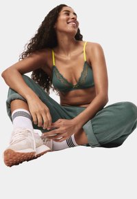 Zielony koronkowy biustonosz z żółtymi ramiączkami, zestawiony z zielonymi spodniami dresowymi. Modelka nosi białe skarpetki z czarnymi paskami i jasne sneakersy.