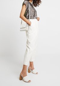Weiße hochtaillierte Hose, gemustertes graues Oberteil, cremefarbener Umhängetasche und weiße Sandalen mit offenem Zeh und Blockabsatz mit geflochtenem Design.