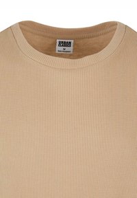 Béžový rebrovaný sveter s okrúhlym výstrihom a presne šitými okrajmi. Nálepka hovorí "URBAN CLASSICS" s veľkosťou "M." Viditeľná jemná textúra.
