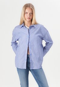Junge Frau in gestreiften blauen Hemd über einem weißen Top und blauen Jeans, die mit Händen hinter dem Rücken vor einem weißen Hintergrund steht.