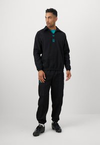 Sort atletisk outfit bestående af en pullover med krave og bukser, med teal logo accenter. Stoffet ser glat ud og har elastiske manchetter.