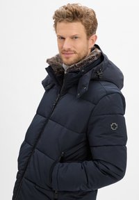 Marine blauwe pufferjacket met een nepbont gevoerde capuchon, ritsluiting, elastische manchetten en zijzakken. Kenmerkend is het logo-op-nu op de mouw.