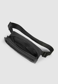 Schwarze kompakte Tasche mit einer strukturierten Außenseite, einem oberen Reißverschluss und einem verstellbaren Riemen. Das Innere ist gefüttert und geräumig, mit Logodetails.