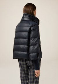 Marineblå pufferjakke med høj krave, quiltet design og blank tekstur. Har sidelommer og en cropped længde.