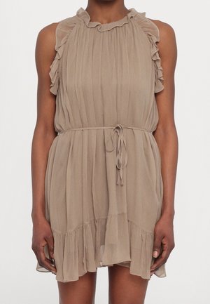 Beżowa sukienka bez rękawów z falbankami na krawędziach, plisowaną tkaniną i cienkim paskiem zawiązanym w talii, noszona przez osobę o ciemnej karnacji.