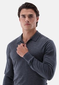 Graues Polohemd mit langen Ärmeln, Kragen, strukturierter Stoff und figurbetonter Schnitt. Das Model hält den Kragen mit einer Hand.