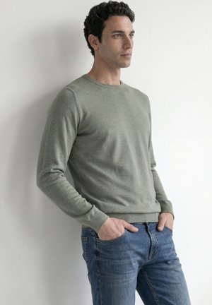 Homme en pull vert et jean bleu, debout les mains dans les poches, regardant sur le côté sur un fond clair uni.