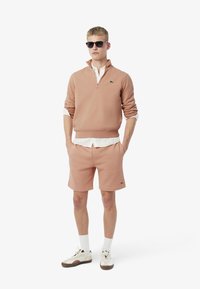 Sudadera rosa claro con cuello de media cremallera, combinada con pantalones cortos a juego. El modelo lleva gafas de sol y una camiseta blanca debajo. Logotipo de cocodrilo notable.