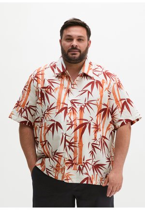 Man with beard wearing short-sleeve white shirt with orange and red bamboo print, standing with one hand in pocket against plain background.