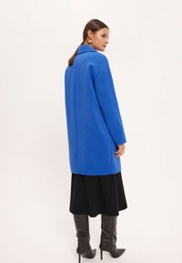 Niebieski, oversize płaszcz z kołnierzem, o sztywnym kroju i długich rękawach, zestawiony z czarną spódnicą i błyszczącymi czarnymi kozakami do kolan.