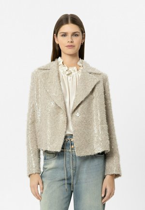 Junge Frau trägt eine beige strukturierte Paillettenjacke über einer cremefarbenen Bluse mit Rüschen und hellblaue hoch taillierte Jeans mit Kordelzug.