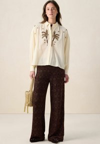 Cream button-up shirt with palm tree and floral embroidery; paired with brown patterned wide-leg pants. Model holds a gold handbag.