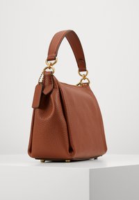 Bolsa de couro castanho com um acabamento texturizado, uma alça curta, detalhes em metal dourado e uma etiqueta pequena anexada, apresentando uma forma estruturada.