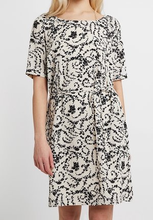 Woman wearing knee-length beige dress with black floral vine patterns and a tied belt at the waist, short sleeves, standing against plain background.