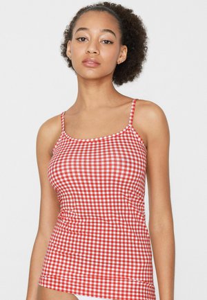 Młoda kobieta o kręconych włosach, ubrana w bezrękawnik w czerwono-białą kratę, na tle jednolitego białego tła.