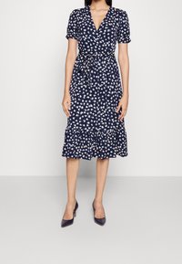 Mekko, jossa on laivastonsinisiä kukkia, lyhyet puffihihat, V-kuvio, kootut vyötärö ja röyhelöhelma; siinä on valkoinen kukkakuosi pehmeällä kankaalla.