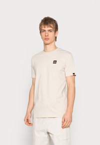 Light beige cotton t-shirt with a crew neckline and short sleeves; features a small black and orange logo patch on the left chest.