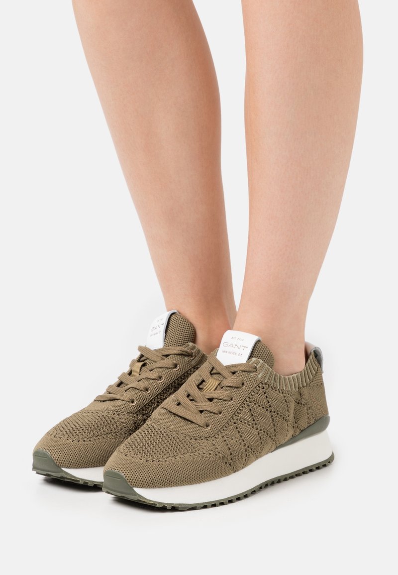 Baskets tricotées vert olive avec semelles et lacets blancs portées pieds nus, avec une étiquette de marque sur la languette et la bride du talon.