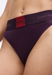 Szilváskék Calvin Klein fehérnemű, kontrasztos piros derékpánttal, dombornyomott logóval, sima textúrával és klasszikus bugyi formával.