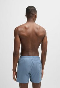 Mies tummalla iholla ja lyhyillä hiuksilla selin kuvattuna, päällä vaaleansiniset uimashortsit, joissa on takatasku, yksinkertaista vaaleaa taustaa vasten.