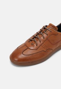 Hnědé kožené tenisky s texturovaným povrchem, šněrovacím designem a jemným červeným prošíváním podél boků a špičky. Gumová podešev.