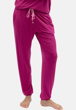 Person, die locker sitzende, knallpinke Freizeit-Hose mit Kordelzug in der Taille und Knöpfen trägt, steht barfuß vor einem einfarbigen Hintergrund.