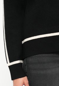 Melnā džemperis ar baltām svītrām, piegrieztnēm un apakšmalu. Redzama mīksta tekstūra, saskaņota ar tumši pelēkiem džinsiem.