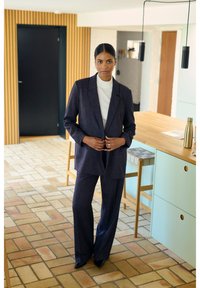Completo su misura blu scuro con giacca lunga e pantaloni a gamba larga. Indossato sopra un dolcevita bianco. Tessuto liscio, design minimalista.