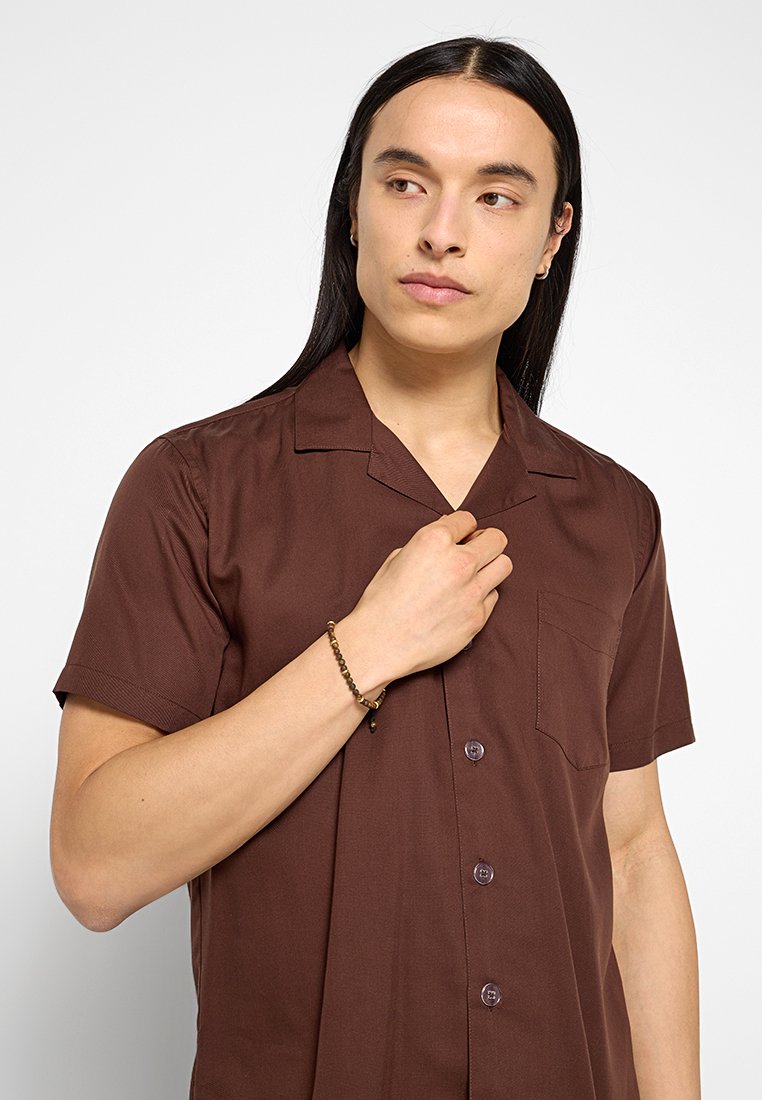 Chemise marron à manches courtes avec un col classique, une poche poitrine unique et un boutonnage à l'avant. Texture lisse et tissu léger.