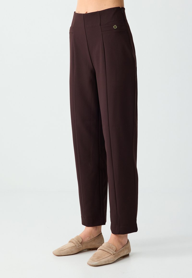Pantalon droit marron foncé avec poches latérales, porté avec des mocassins en daim beige sur un fond clair.