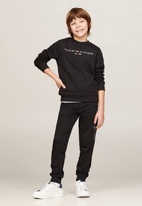 Felpa nera con testo bianco e rosso, abbinata a pantaloni jogger neri con logo. Sneakers bianche con accenti blu e rossi.