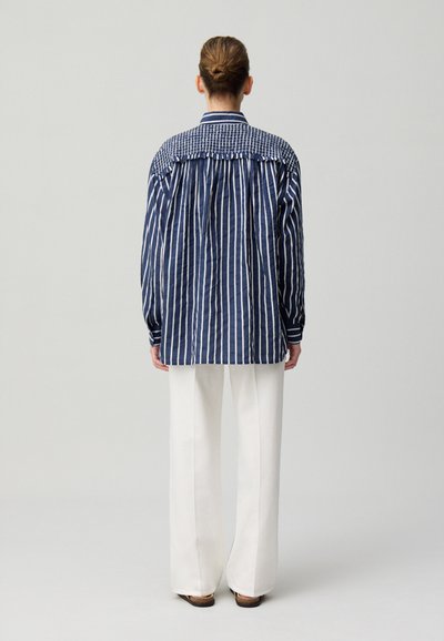 Camisa de botones a rayas en azul marino y blanco, con una pechera con patrón, mangas largas y un corte relajado, combinada con unos pantalones de pierna ancha blancos.