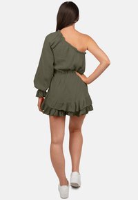 Robe vert olive à une épaule avec une longue manche, taille froncée et ourlet à volants. Fabriquée en tissu texturé. Portée avec des baskets blanches.