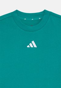 Turquoise t-shirt met korte mouwen, een geribde ronde hals en een wit Adidas-logo op de voorkant. Het materiaal is zacht en glad.