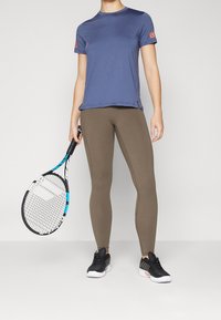 Blå idrottströja med orange logotyp, olivgröna leggings och svarta sportskor. Håller en tennisracket med blå detaljer.