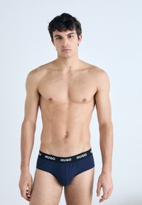 Nachtblaues Baumwollunterwäsche mit einem schwarzen Bund, der mit weißem "HUGO"-Branding versehen ist, präsentiert auf einem männlichen Modell vor einem neutralen Hintergrund.