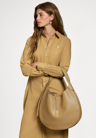 Torba hobo od smeđe kože oblikovana u zakrivljeni oblik, glatke teksture i sa zlatnim detaljem, nošena preko ramena s bež haljinom na kopčanje.
