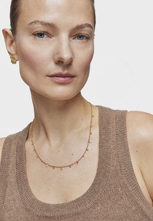 Femme portant des boucles d'oreilles clous en or, un collier délicat en chaîne d'or avec de petits pendentifs, et un haut sans manches en maille marron sur un fond uni.