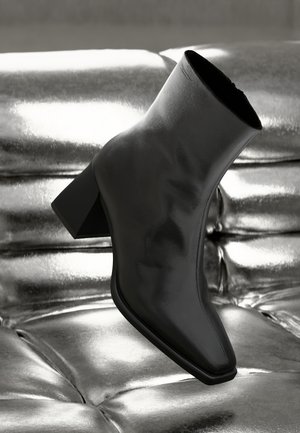 Zwarte leren enkellaars met blokhak geplaatst op een glanzend, metallisch getufte oppervlak in monochrome verlichting.