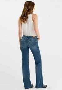 Jeans bleu évasés avec une taille mi-haute, dotés de deux poches arrière avec une couture en zigzag subtile et d'une plaque en cuir beige au niveau de la taille.