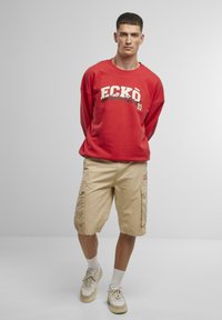 Felpa rossa con grafica "ECKŌ" e pantaloni cargo khaki. Abbinata a calzini bianchi e sneakers beige. Sfondo semplice.