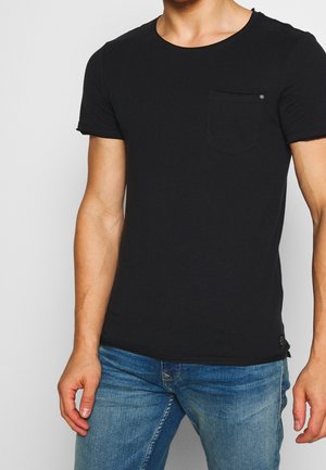 Homme portant un T-shirt noir uni à manches courtes avec une petite poche sur la poitrine et un jean bleu, sur un fond neutre.