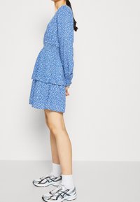Robe bleue à pois blancs, manches longues, design à volants, taille élastique. Portée avec des chaussettes blanches et des chaussures de sport multicolores.