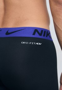 Leggings Dri-FIT noirs avec une finition texturée, présentant une taille violette et un logo Nike noir audacieux. Branding Dri-FIT ADV visible.
